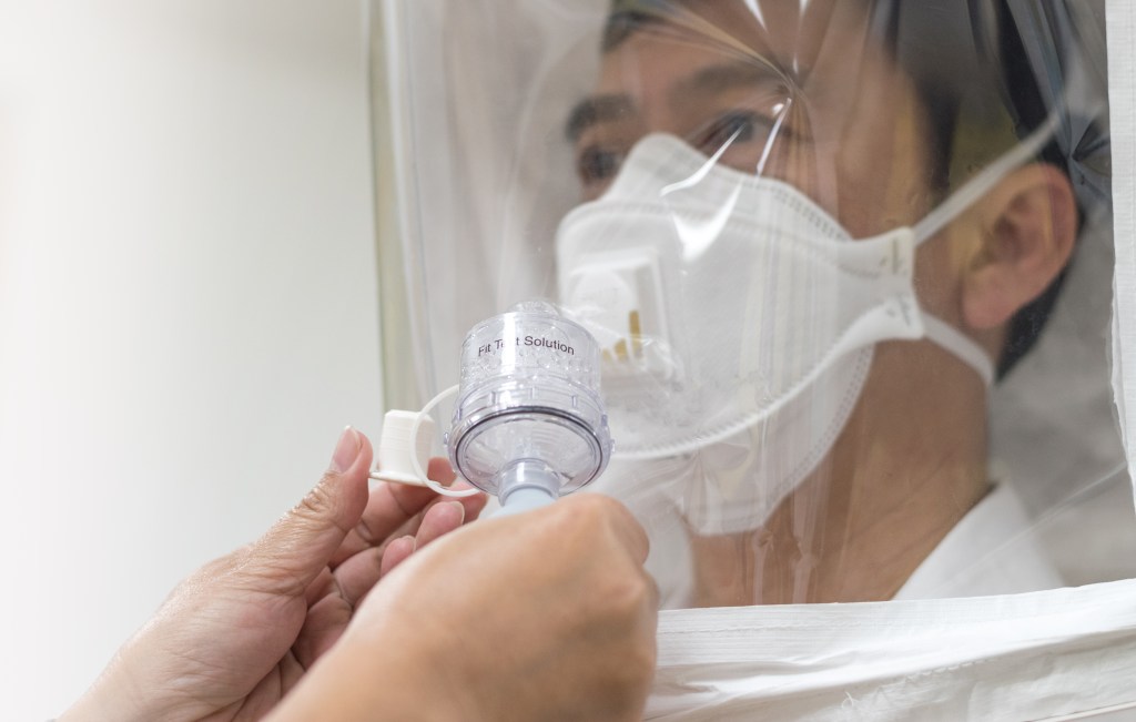 An man is wearing an N-95 mask. He has a white hood over his head where a bitter tasting solution is being fed into the hood to conduct a qualitative respirator fit test.